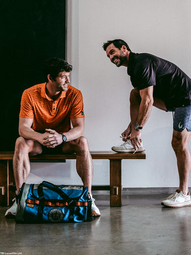 Two men in a casual setting with one sitting on a bench and the other standing, near a blue star wars duffel bag.
