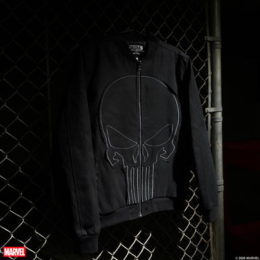 The Punisher Themed Zip Up Bomber Jacket with Black Monochromatic patch and contrast white stitching on a dark background hanging on a metal fence