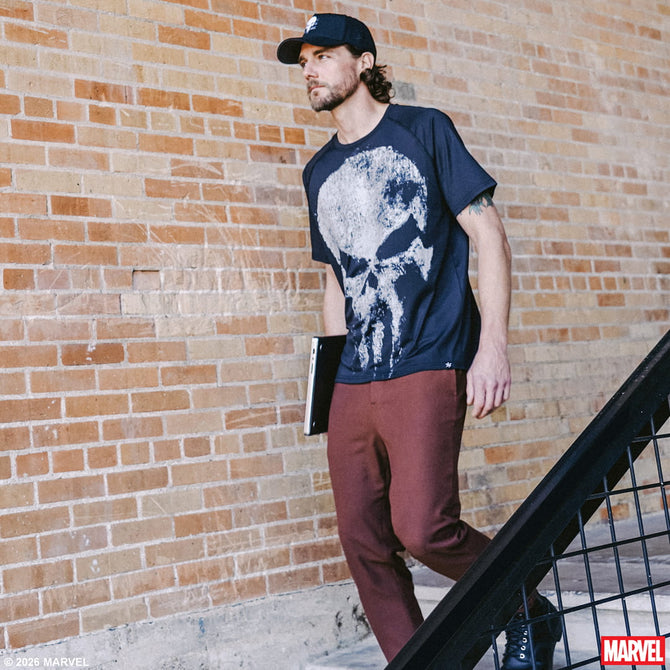 Man wearing a Punisher t-shirt with a skull design, maroon pants, and a black cap against a brick wall.