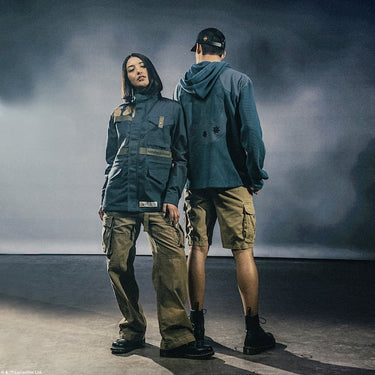 Two people wearing teal green Star Wars Rebel Pathfinders jackets and brown pants standing against a dark, smoky background.
