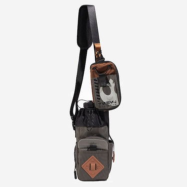 The photo shows a crossbody bag designed for carrying a water bottle. The bag features a drawstring closure at the top, a front zippered pocket, and an orange rubber patch with a geometric design meant for attaching a clip. The adjustable black strap has a padded section for comfort and a blue cord with holes to attach clips and a tech organizer bag. The bag is in a subtle brown with pops of orange and black.