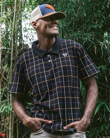 A man standing outdoors wearing a plaid superman themed polo and a superman themed trucker hat