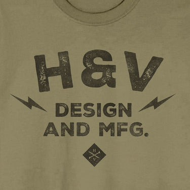 The photo shows a close up of a shirts print. The print on the shirt says "H&V  Design and MFG." The text is stacked with "H&V" on top, "Design" in the middle and "And MFG." below. On either side of the word "design" is a small lightening bolt shape. Under the  text "And MFG" is a small diamond shape with a cross bones logo. 