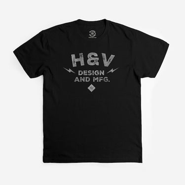 The photo shows a black short sleeve shirt. The print on the shirt says "H&V   Design and MFG." The text is stacked with "H&V" on top, "Design" in the middle and "And MFG." below. On either side of the word "design" is a small lightening bolt shape. Under the  text "And MFG" is a small diamond shape with a cross bones logo. 