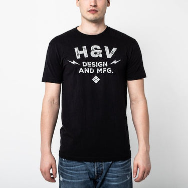The photo shows a man in a black short sleeve shirt. The print on the shirt says "H&V   Design and MFG." The text is stacked with "H&V" on top, "Design" in the middle and "And MFG." below. On either side of the word "design" is a small lightening bolt shape. Under the  text "And MFG" is a small diamond shape with a cross bones logo. 