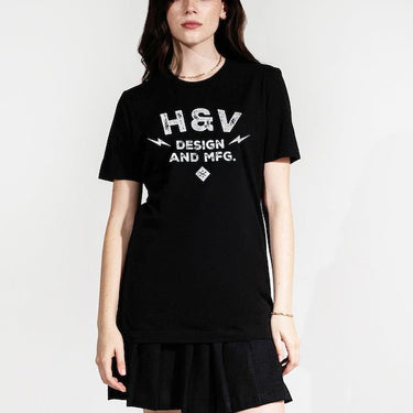 The photo shows a woman in a black short sleeve shirt. The print on the shirt says "H&V   Design and MFG." The text is stacked with "H&V" on top, "Design" in the middle and "And MFG." below. On either side of the word "design" is a small lightening bolt shape. Under the  text "And MFG" is a small diamond shape with a cross bones logo. 
