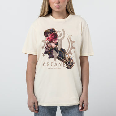 A female model wearing a cream-colored Arcane x League of Legends t-shirt featuring Vi in an action pose. She has her signature pink hair and wields her massive Hextech gauntlets, detailed with metallic and glowing blue accents. Behind her, a faded Piltover gear emblem adds depth to the design. 'Arcane' and 'League of Legends' are printed below in a bold, distressed font