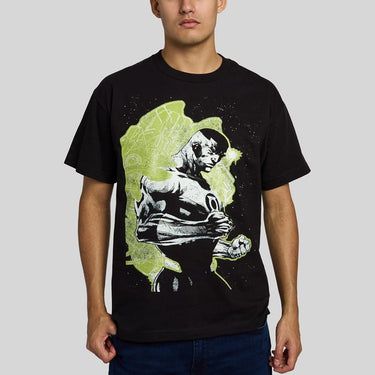 A man wearing a black short-sleeve graphic t-shirt with a stylized print of Green Lantern, set against a lime green and black background, paired with dark blue jeans, on a solid gray studio background.