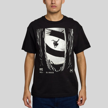 A man wearing A black short-sleeve crewneck t-shirt featuring a large black and white graphic print of Spider-Man swinging from a web on a striped banner, with 'NYC WEB SLINGER' text printed below the graphic, paired with dark blue jeans, against a solid gray background.