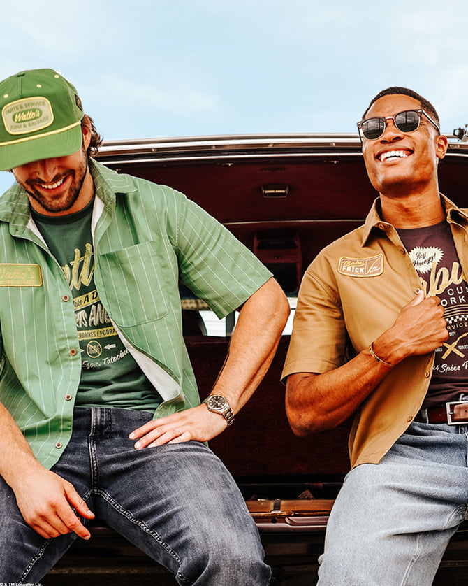 Two men smiling and posing together in front of the open trunk of a vintage car. The man on the left wears a green cap, a green striped button-up shirt over a dark graphic tee, and jeans.